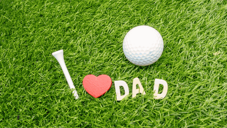 A golf ball, golf tee, and wooden letters spelling “I ♥ DAD” arranged on bright green grass, representing a Father’s Day golf gift or celebration.